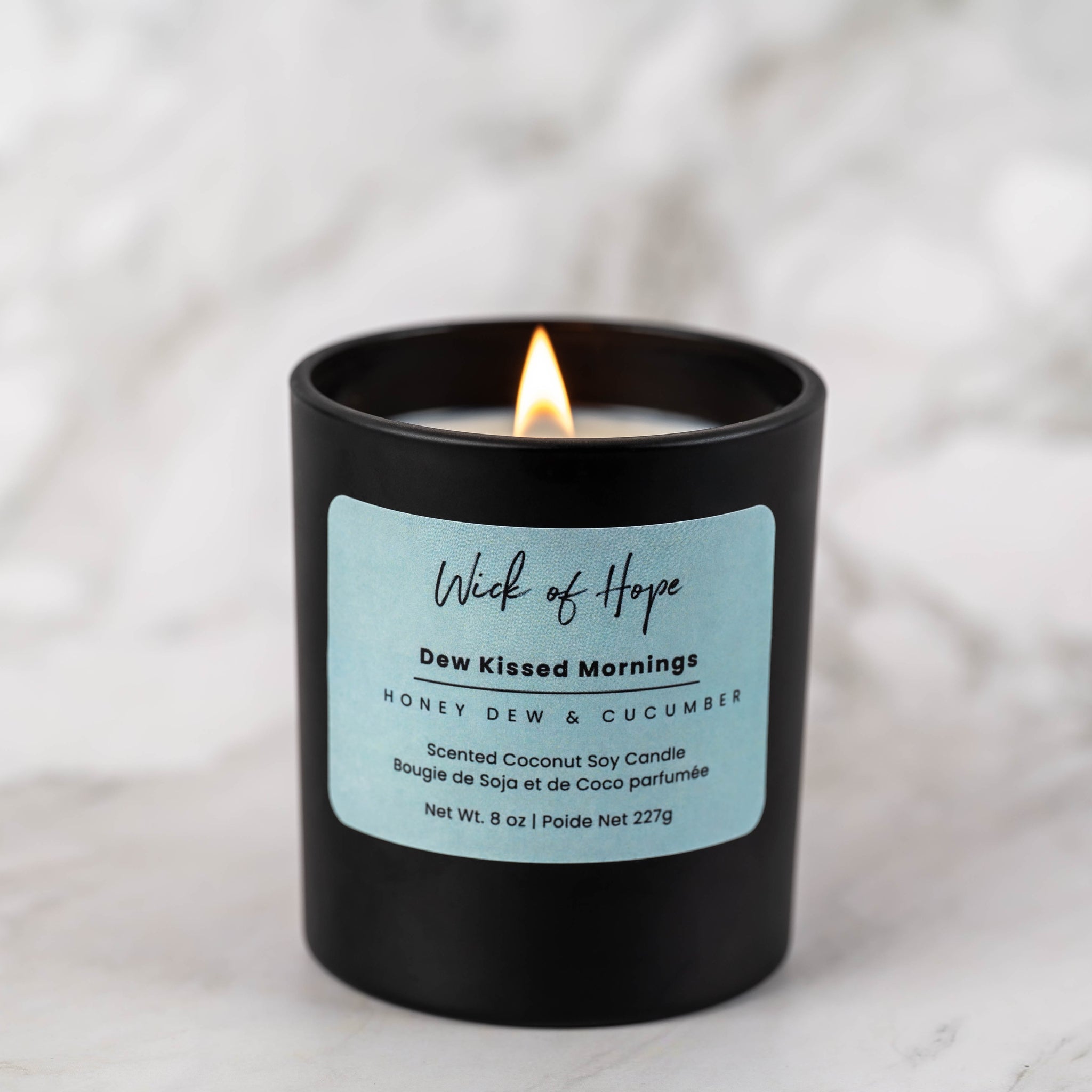 A black candle labeled Wick of Hope with a light blue sticker and burning flame sits on a white marble surface. The label reads Dew Kissed Mornings, Honey Dew & Cucumber, Scented Coconut Soy Candle.